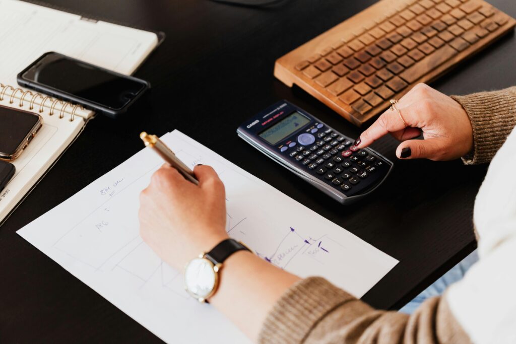A desk with a notebook, calculator and keyboard with someone writing and tapping on the calculator. Likely, the person could do with some financial advice or support