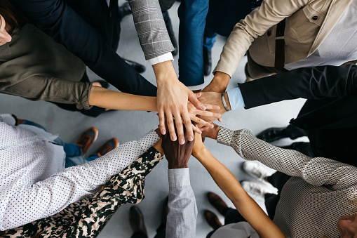 A high-angle shot of a group of male and female colleagues putting their hands together in an office demonstrating team work and building a team. Their faces are not visible, only their arms.