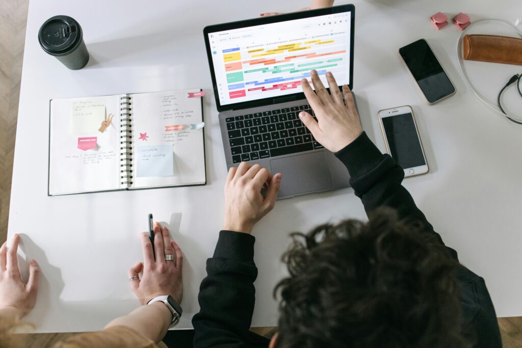 Overhead view of two people collaborating on a content creation service project. One person is pointing at a colorful project timeline on a laptop screen, while the other is taking notes in an open notebook filled with sticky notes and task lists. A smartphone, coffee cup, and other office supplies are spread across the white desk, suggesting a productive virtual assistant content creation service.