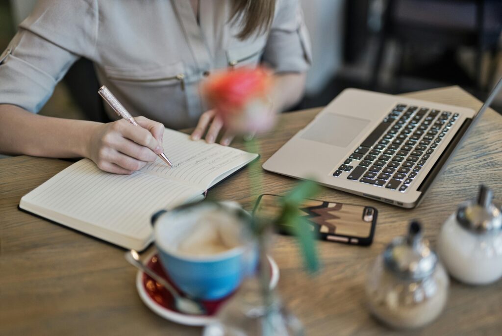 Close-up of a person writing in a notebook next to an open laptop and a coffee cup on a table, with a blurred flower in the foreground. The scene conveys organisation and focus. How to set your week up for success.