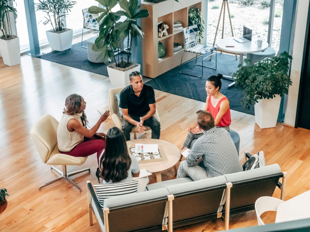 A diverse group of five people engaged in a casual, collaborative discussion around a small round table in a modern office space. The environment features plants, large windows, and comfortable seating, reflecting a company culture that promotes teamwork, inclusivity, and open communication, emphasising the importance of culture fit within the team.