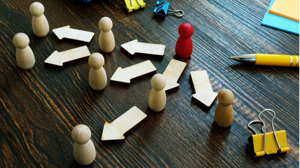 Wooden figurines with arrows connecting them on a desk, representing delegation and task flow. A single red figure stands out, symbolising a bottleneck or inefficiency in the process. This visual illustrates how ineffective delegation leads to missed opportunity cost in business and teamwork.